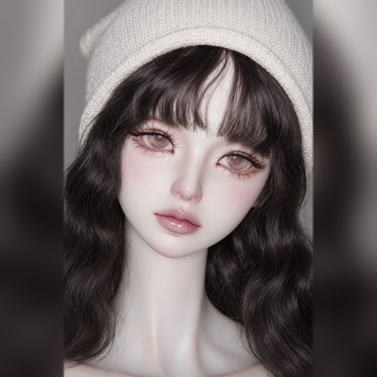 Three-quarter close-up of a female BJD head with delicate pink face-up, long dark waves and a cream knit beanie