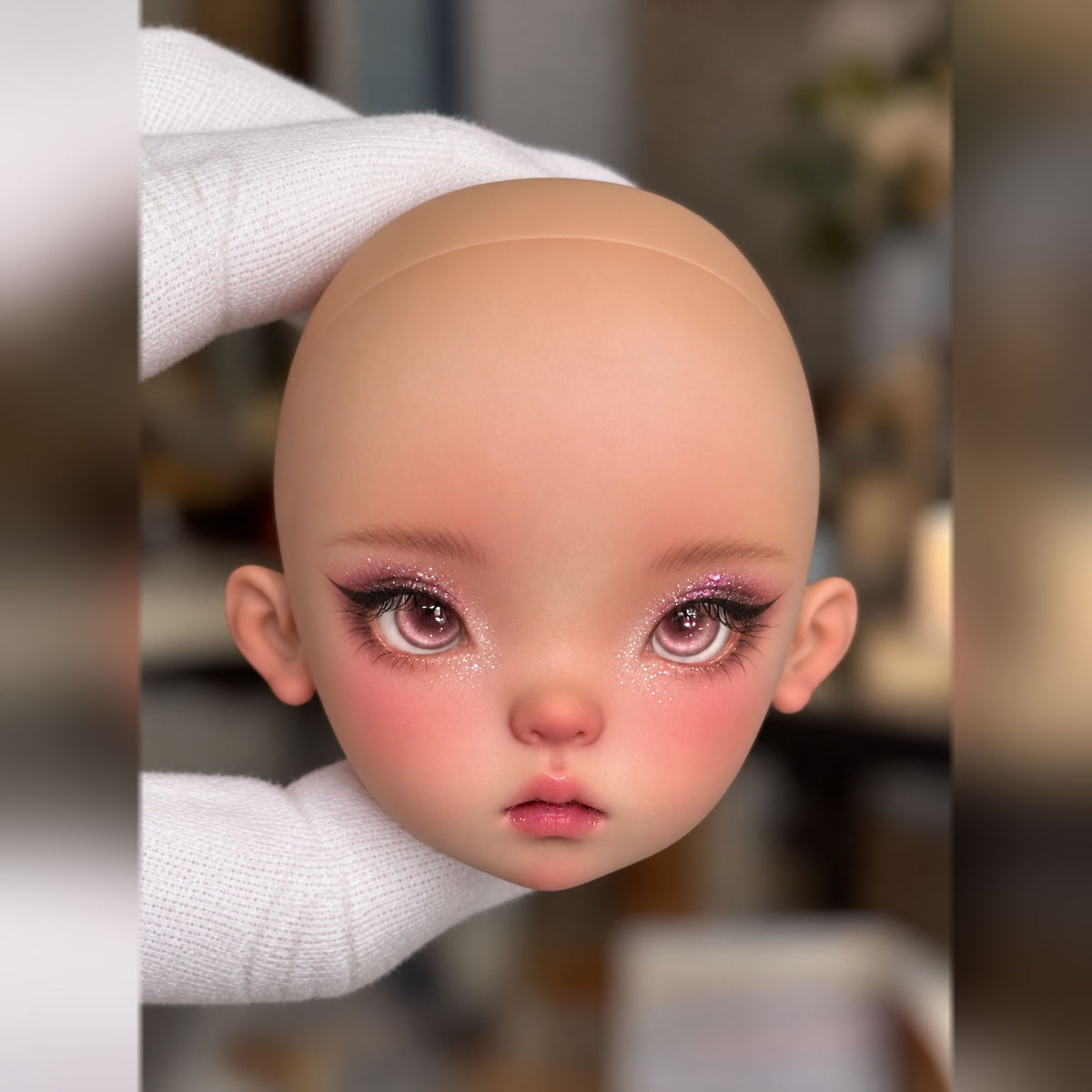 Front view of a made-up BJD head with large pink eyes, sparkling eye makeup, rosy blush and a small open mouth
