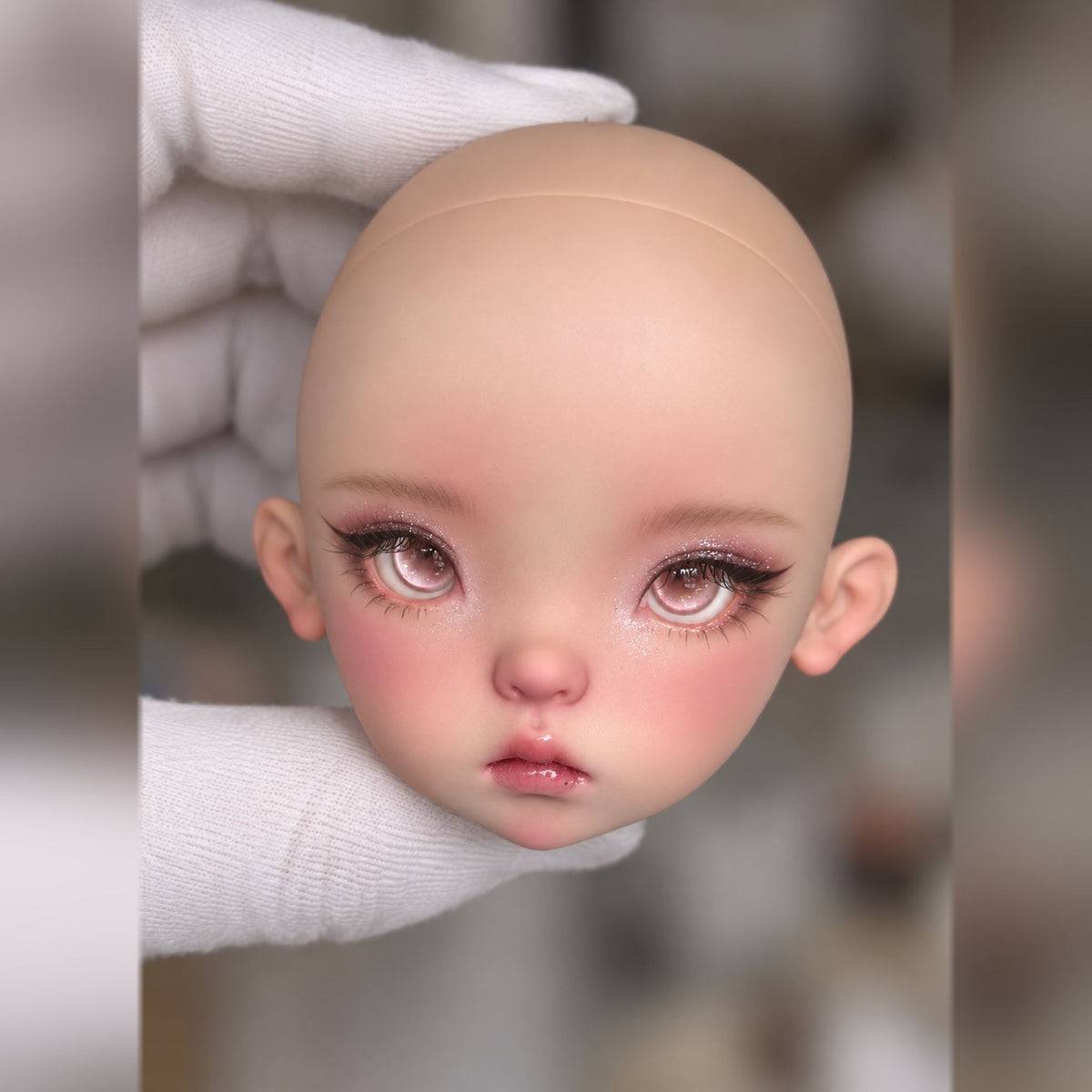 Three-quarter view of a made-up resin doll head with pink eyes, long lashes, glitter accents and glossy lips
